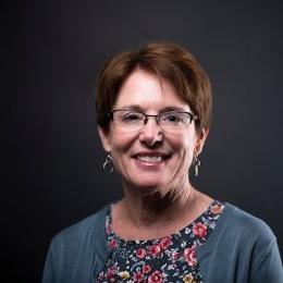 Lady with short brown hair wearing grey cardigan and multi flowered top and glasses in front of grey background