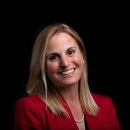 Lady with shoulder length blonde hair with red blazer and pearls in front of black background