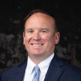 Man with dark suit wearing light blue tie and white button up in front of grey background