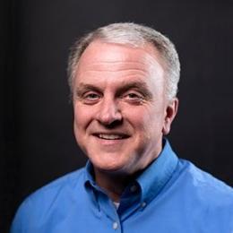 Man with short grey hair with blue button up dress shirt in front of black background