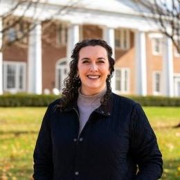 Lady with dark curly hair wearing dark jacket and lavender turtleneck in front of Old Centre