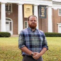 Man with short auburn hair and beard wearing plaid button up shirt standing in front of Old Centre