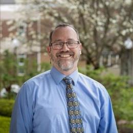 Man with dark hair and beard wearing glasses, earring on left ear, blue button up dress shirt and tie