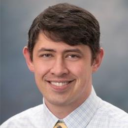 Man with dark hair wearing striped button up dress shirt and yellow tie in front of grey background