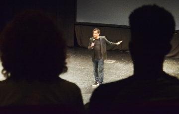 A man holding a microphone is seen between the silhouettes of two audience members. 