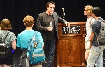 Students carrying backpacks stand in a loose circle around a man holding a water bottle and resting his arm on a podium bearing the 中央书院标志. 