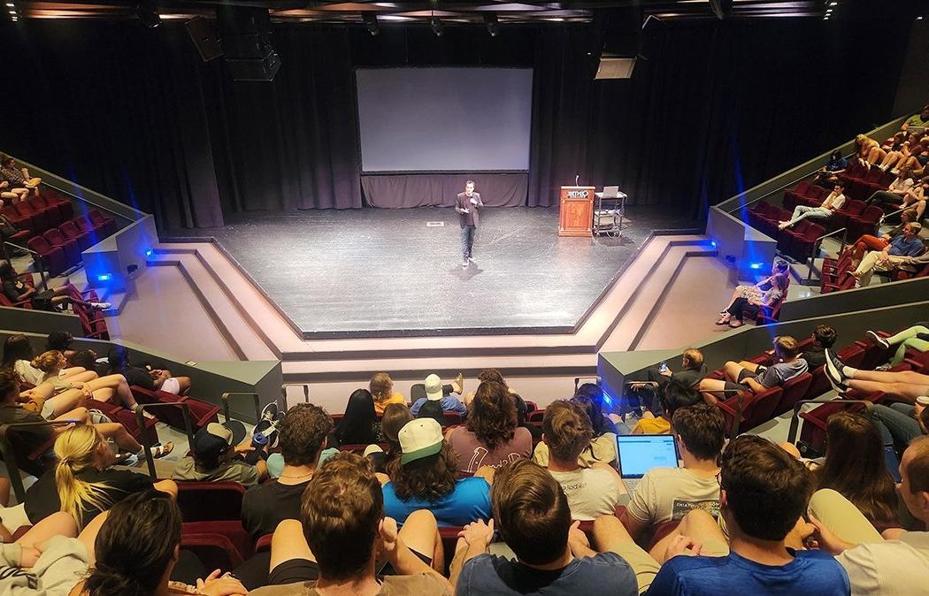 A man talks into a microphone while standing on a stage surrounded by theatre seats filled with students. 